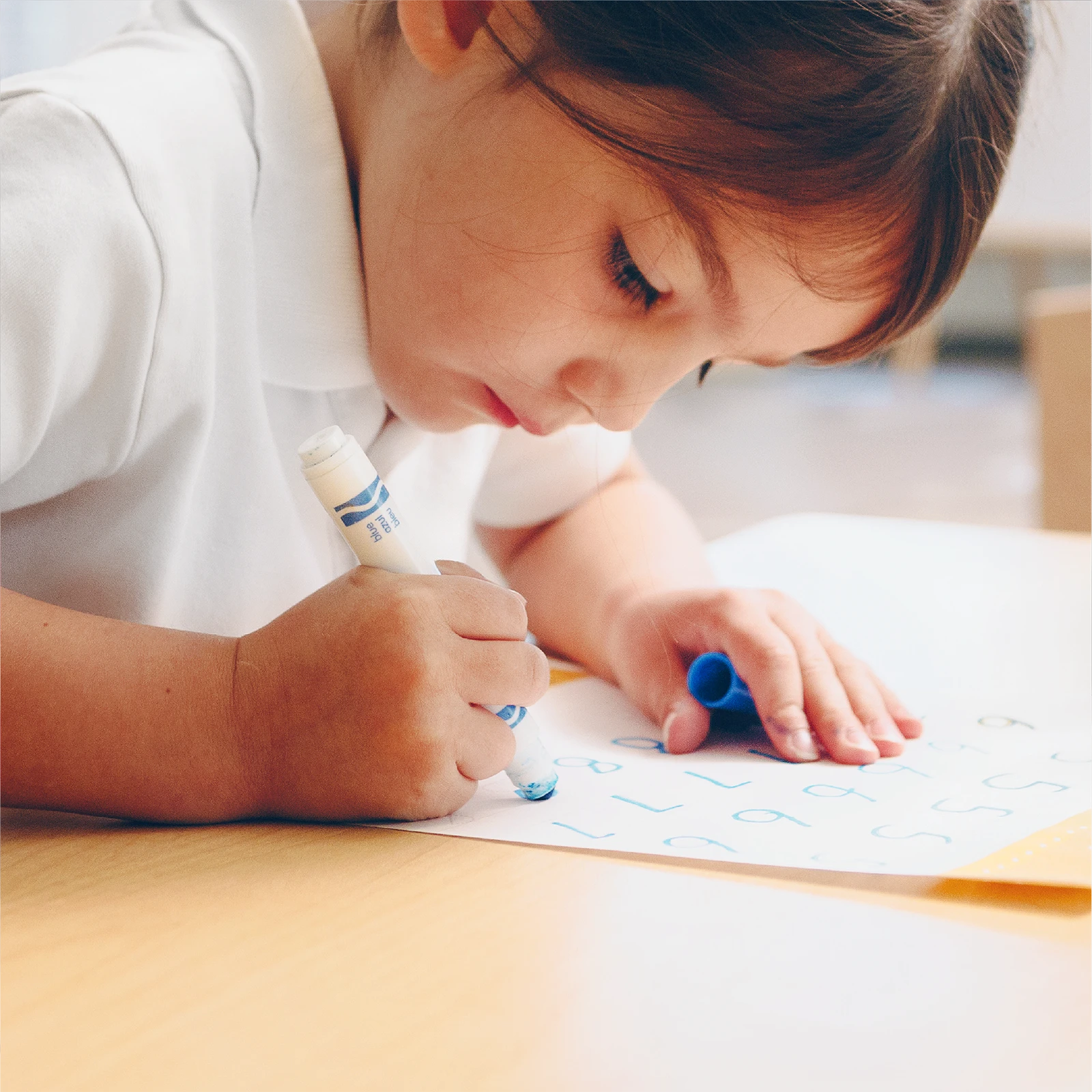 Young child in a white shirt uses a blue marker to trace letters on a sheet of paper while sitting at a table, focused on practicing writing skills, much like activities found in early learning centres.