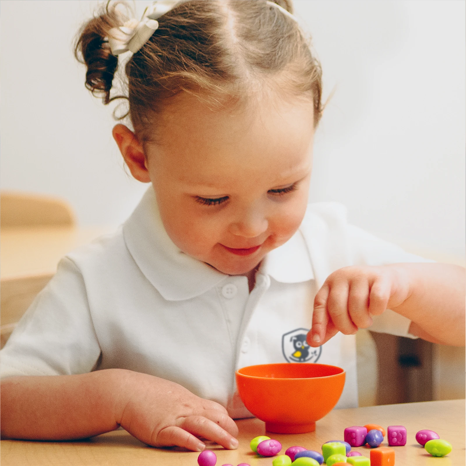 A young child with light brown hair in pigtails sits at a table, smiling and sorting colorful beads into an orange bowl, enjoying hands-on activities common in early learning centres.