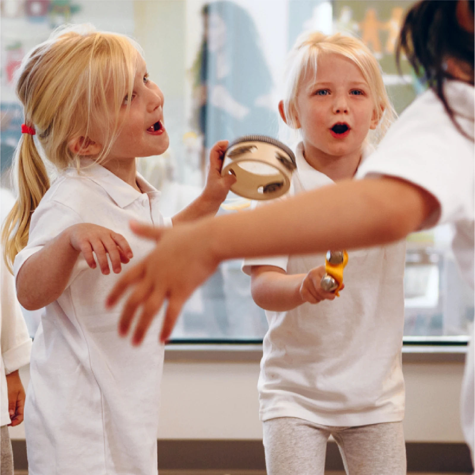 Two young girls in white shirts enthusiastically play musical instruments, a tambourine and maracas, while singing indoors at an early learning centre. Another child's outstretched arm is visible in the foreground.