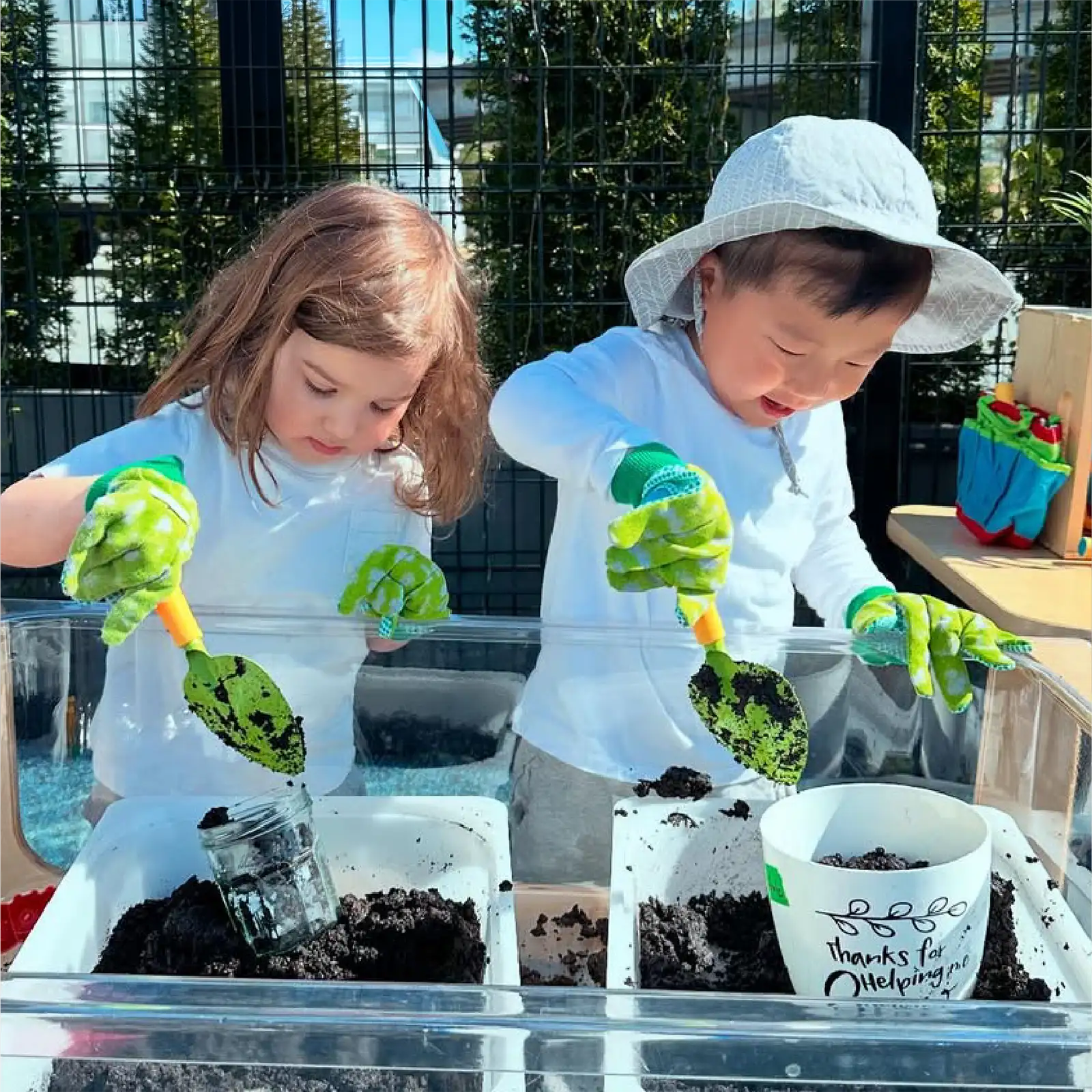 Two children wearing gloves dig in soil with small shovels at a clear outdoor planting table. They are focused on their gardening activity, which is part of the hands-on experiences offered by early learning centres.