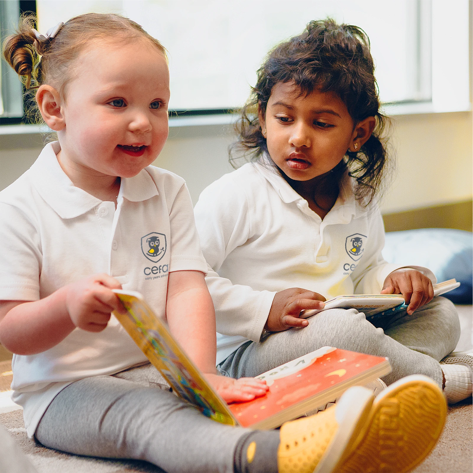 Two young children in white polo shirts and grey pants sit on the floor, holding colorful books. One smiles and talks while the other listens attentively—capturing a joyful moment at one of the top early learning centres.