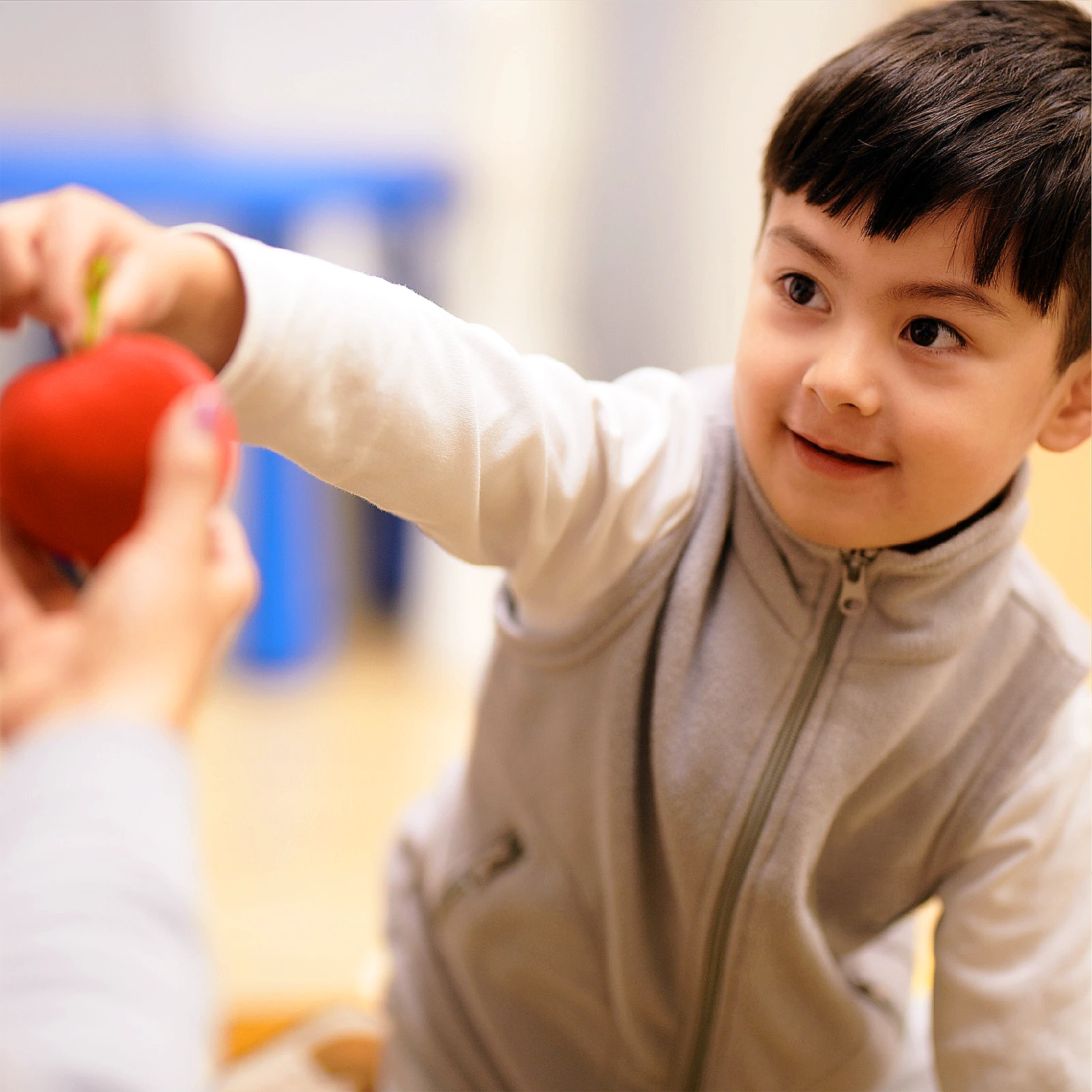 A young child with dark hair and a light gray vest reaches out with a smile to take a red apple from someone’s hand, capturing the joyful spirit found in early learning centres.