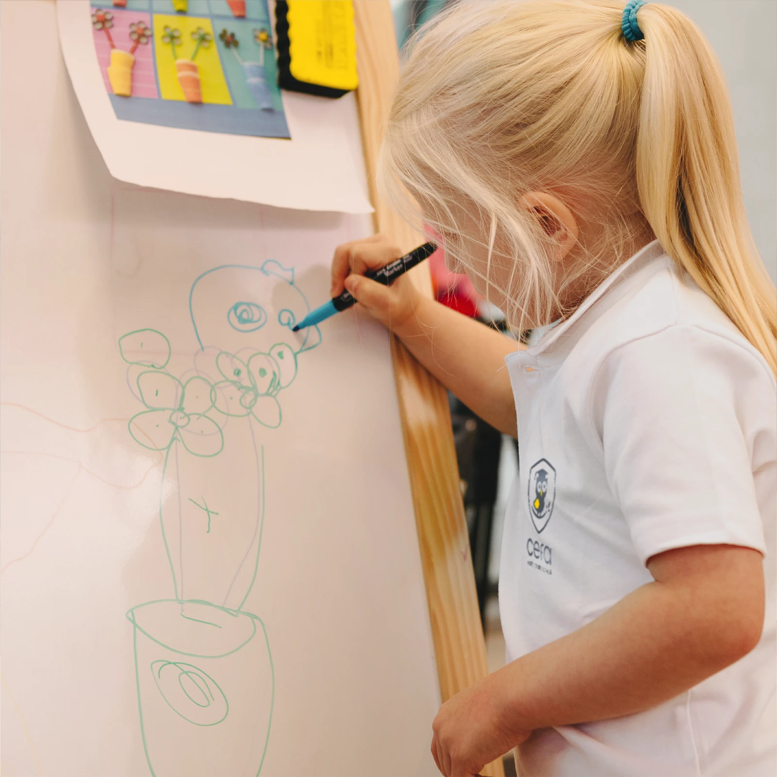 A young child with blonde hair draws colorful flowers on a whiteboard at one of the early learning centres, wearing a white polo with a school logo. A reference photo of flowers is clipped to the top of the board.