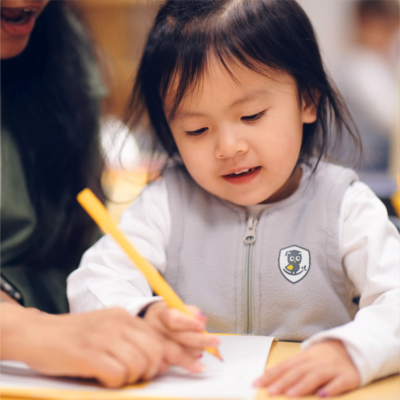 A young child with long dark hair smiles while writing with a yellow pencil, guided by an adult’s hand—showcasing the nurturing environment found in quality early learning centres and early childhood care programs.