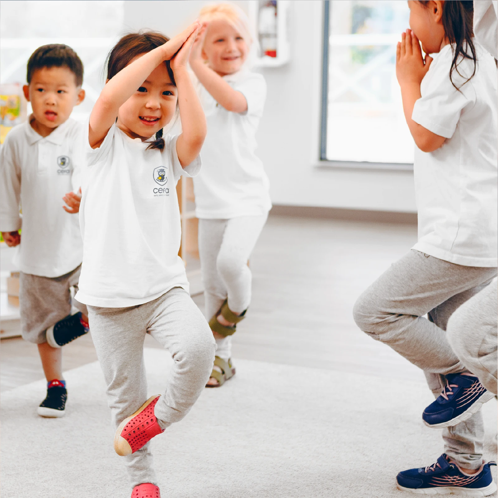 Young children in white shirts and gray pants practice a yoga pose indoors at early learning centres, standing on one leg with hands pressed together above their heads, smiling and appearing joyful.